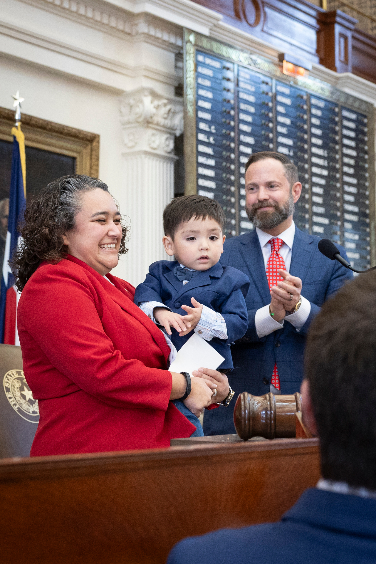 Denise in her red blazer holding Zeke at the dais of the Texas House.